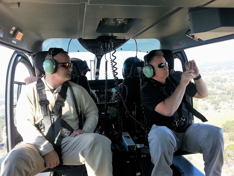 Two men in the helicopter, mid-flight, one is taking a picture of the scenery on his phone.